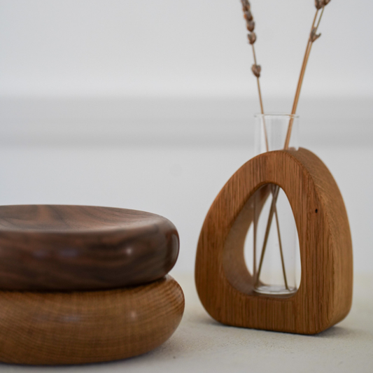 Two wood pillows stacked next to a wood bud vase in a pebble shape with a glass insert holding dried plants, on a neutral background.