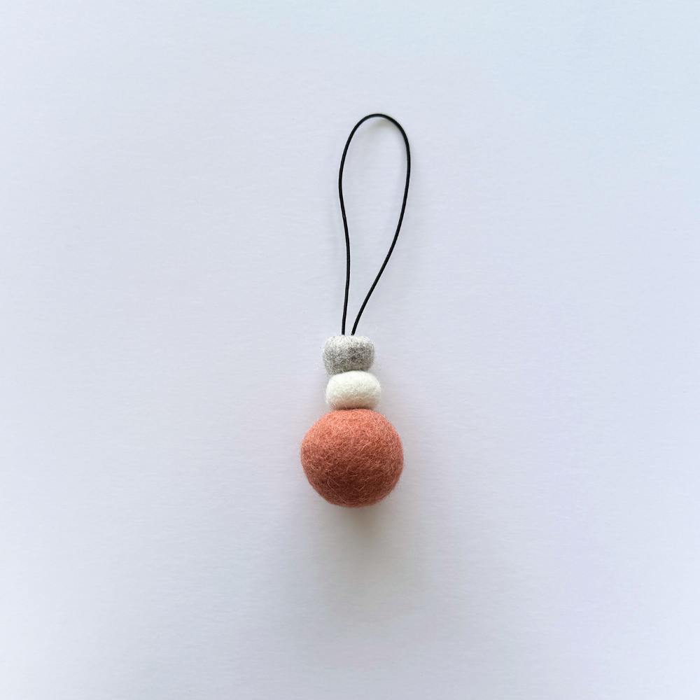 Hand-felted wool hanging diffuser in soft sunset pink with neutral accent beads and black loop, photographed on a light background. Ideal for essential oils in car or home.