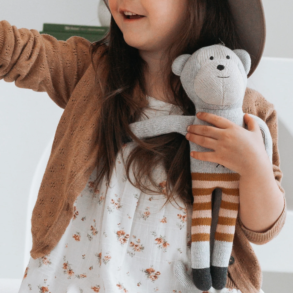 Child holding a monkey soft toy wearing a brown sweater and floral dress.