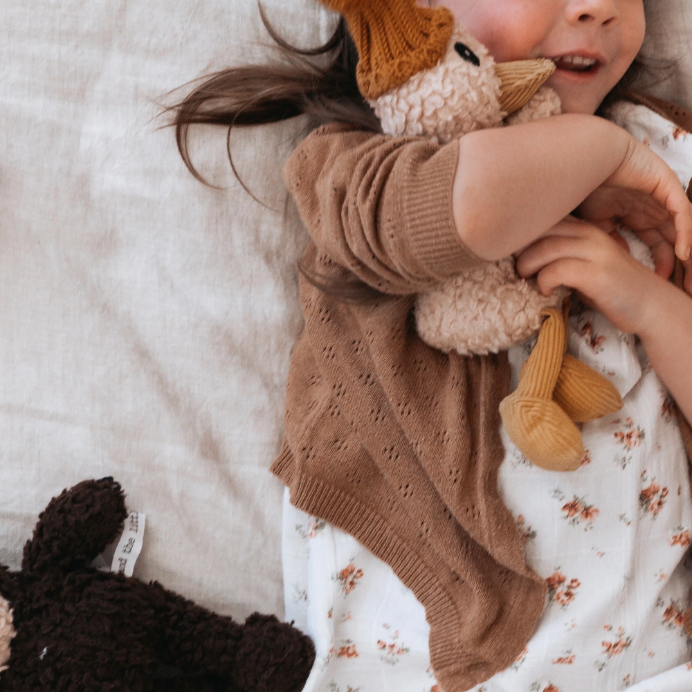 Child wearing a brown knitted sweater holding a duck plush toy on a soft surface.