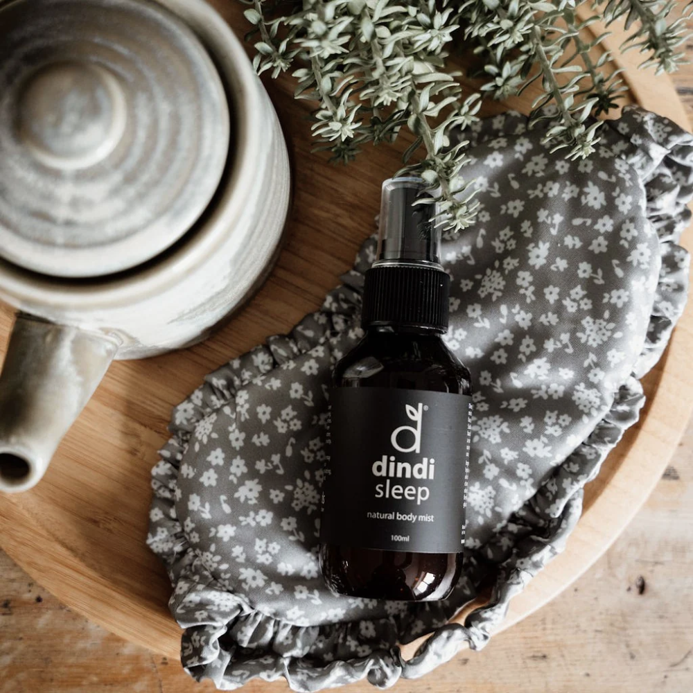 Dindi Sleep natural body mist bottle on a floral-patterned cloth with a ceramic mug and greenery in the background.