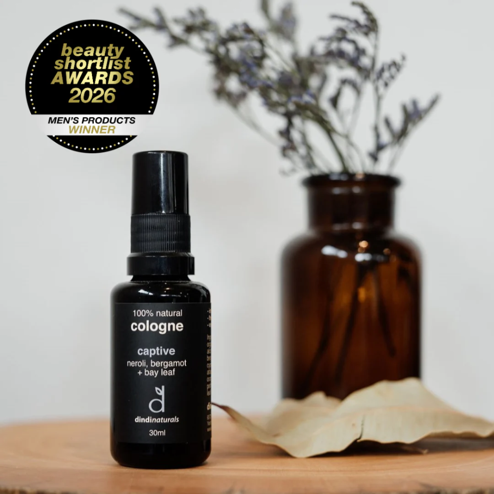 Black cologne bottle with 'Dindi Naturals' branding on a wooden surface, with a vase of dried flowers in the background.
