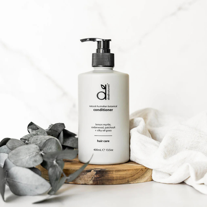 White bottle of conditioner with pump on a wooden board with eucalyptus leaves and a white cloth in the background.