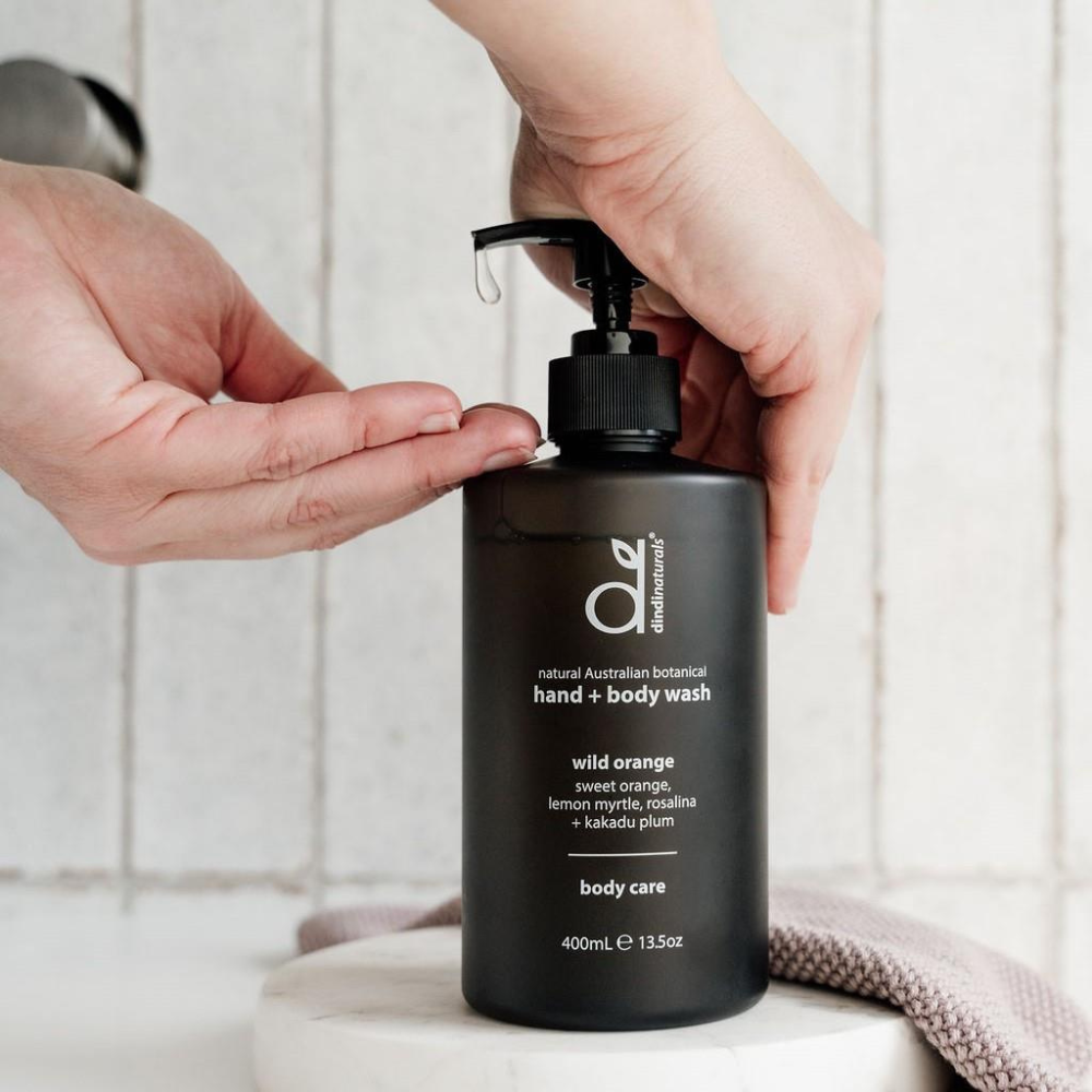 Person holding a black bottle of hand and body wash with a pump, against a tiled bathroom wall.