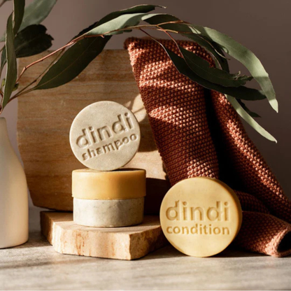 Dindi Naturals Shampoo and Conditioner Bars on a stone block with a towel and plant in the background
