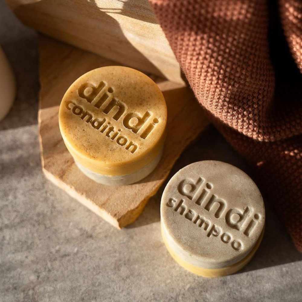 Two Dindi Naturals shampoo and conditioner bars on a wooden stand with a towel in the background.