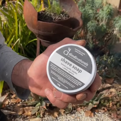 Hand holding a container of Dindi Naturals shave soap outdoors.