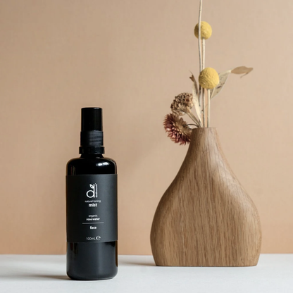Black bottle of Dindi Naturals Rose Toning Mist next to a wood vase with dried flowers on a beige background