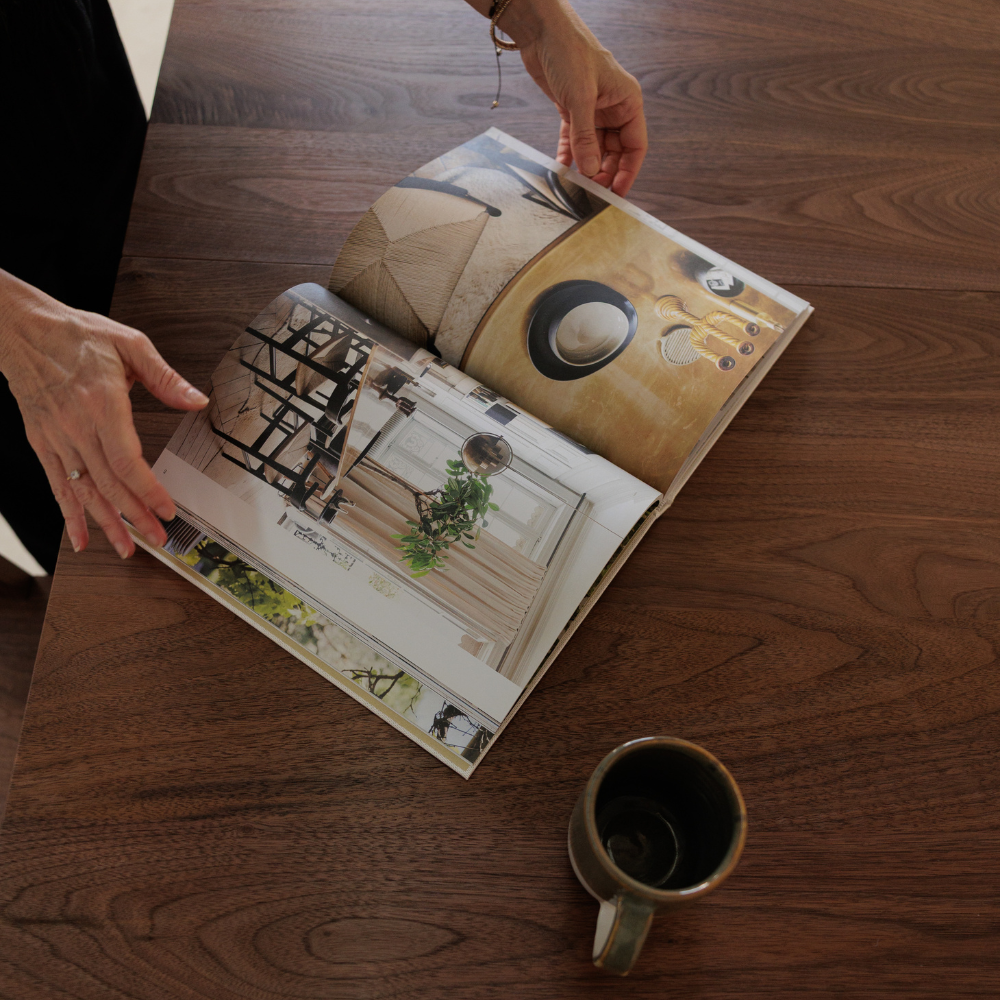 Person flipping through a design book next to a Freya handmade ceramic mug on a wood table.