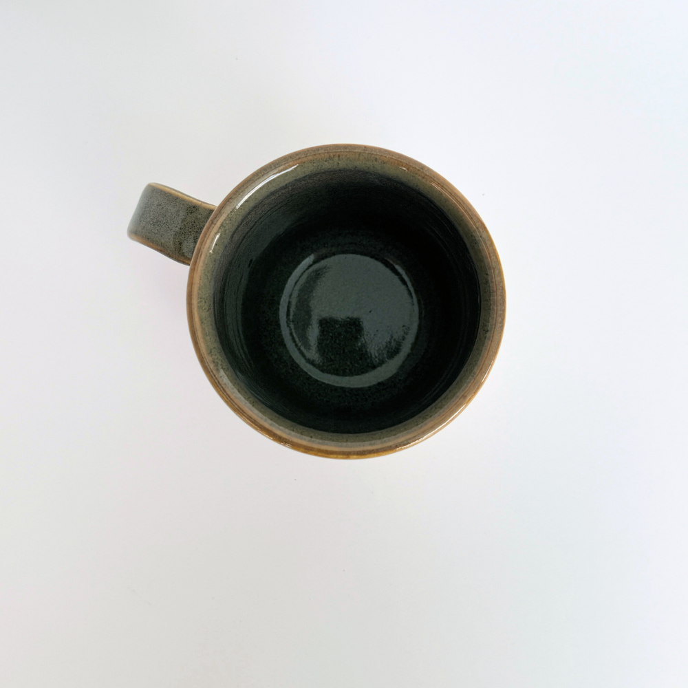 Handmade ceramic mug by artisan Eli C. featuring an earthy green, ochre, and white glaze, displayed on a light backdrop with natural lighting.