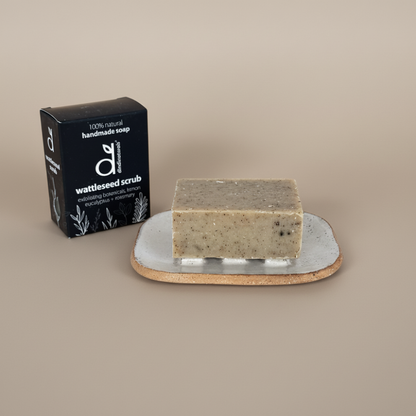 Bar of soap on a soap dish with a black box labelled 'wattleseed scrub' on a beige background