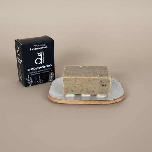 Bar of soap on a soap dish with a black box labelled 'wattleseed scrub' on a beige background
