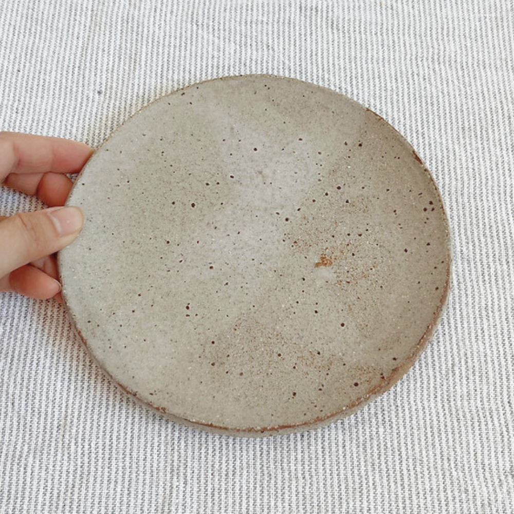 Hand holding a round ceramic plate with a textured surface on a light fabric background