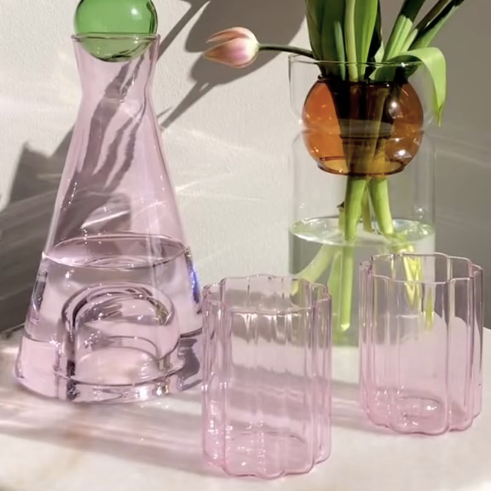 Two pink wave glasses next to a pink glass carafe and a glass vase