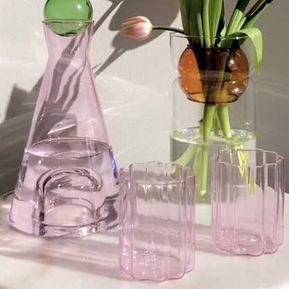 Two pink wave glasses next to a pink glass carafe and a glass vase