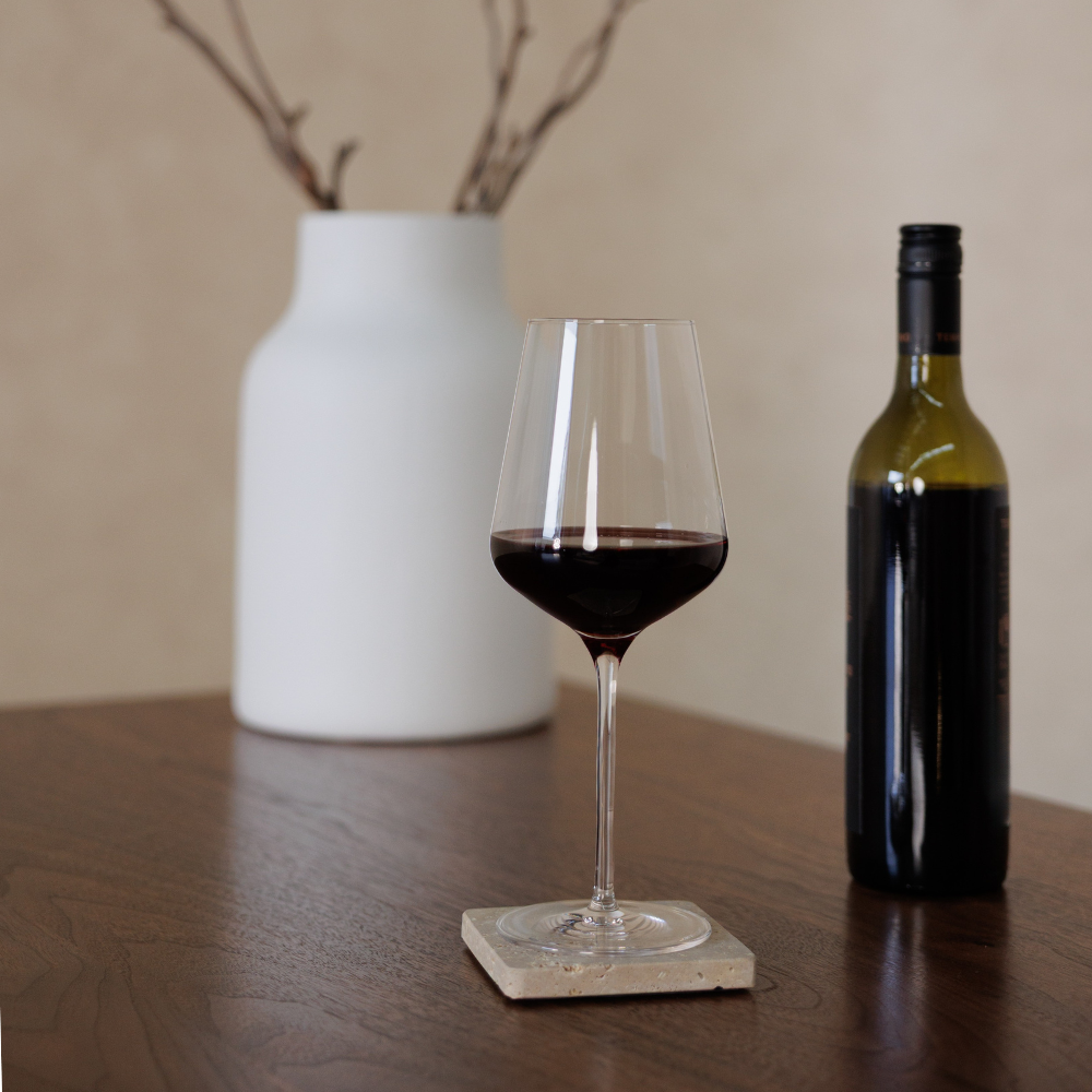 Wine glass with red wine on a travertine coaster next to a bottle of wine and a white vase on a wooden table.