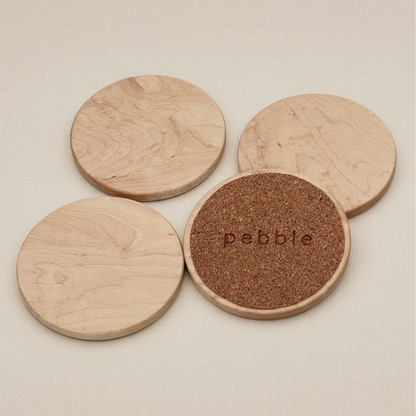 Four wooden coasters with cork bottom on a beige background, one coaster has 'pebble' branding.