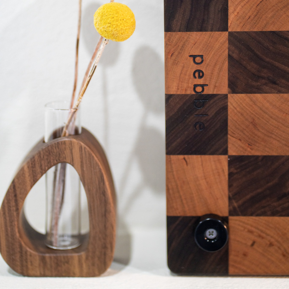 Wooden vase with a yellow flower and checkered wooden board with 'pebble' branding.