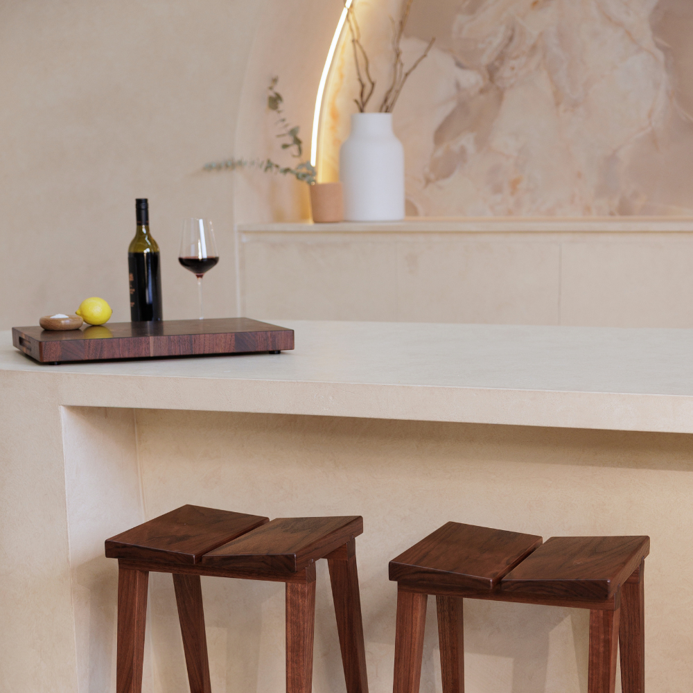 Two wooden stools in front of a bar with a timber chopping board, a bottle and glass.