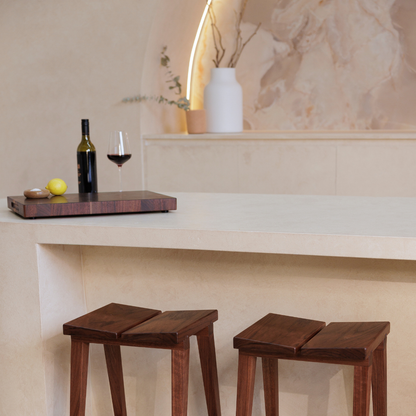 Two wooden stools in front of a bar with a timber chopping board, a bottle and glass.