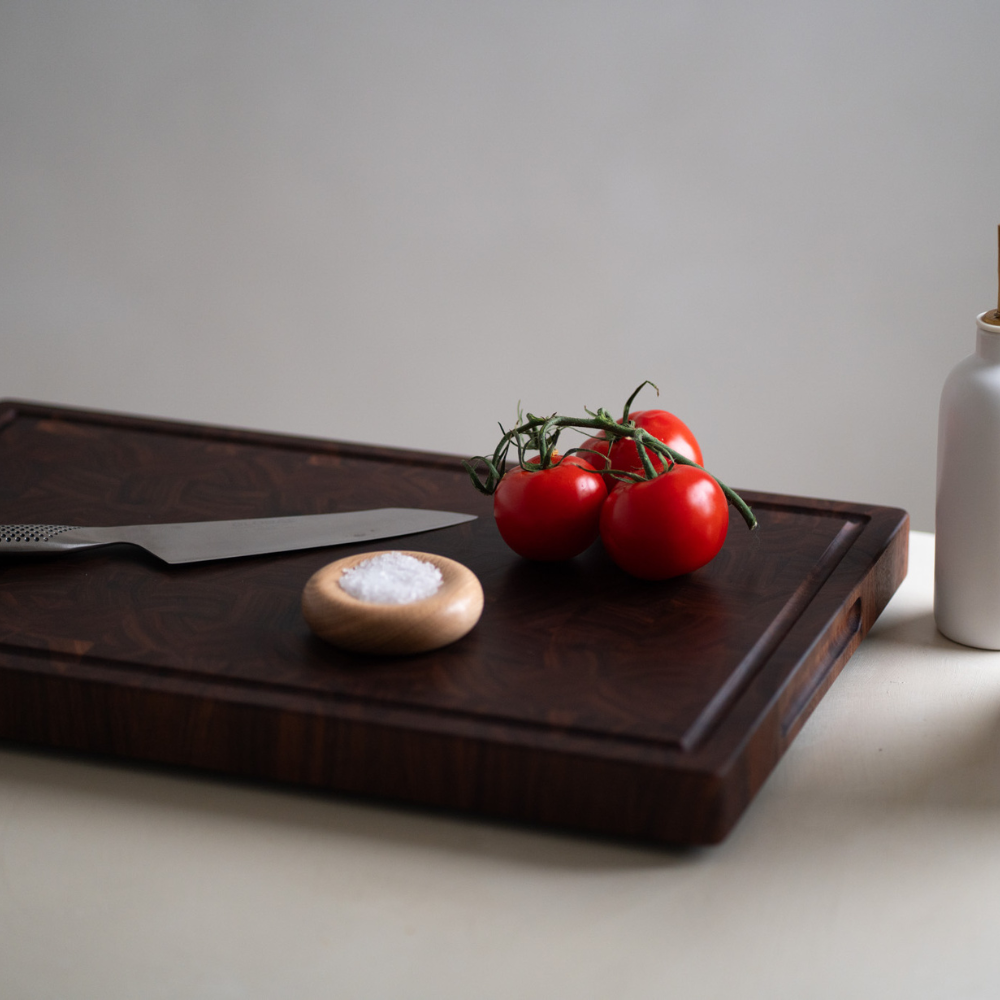 Wooden cutting board with tomatoes, a knife, and a salt container on a neutral background