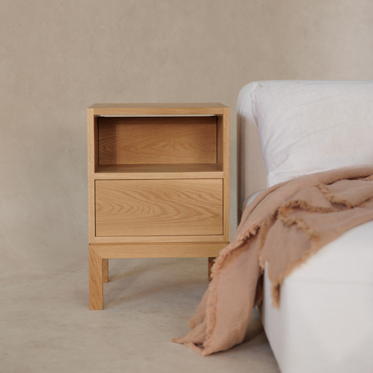Wooden bedside table next to a bed with a beige blanket on a neutral background