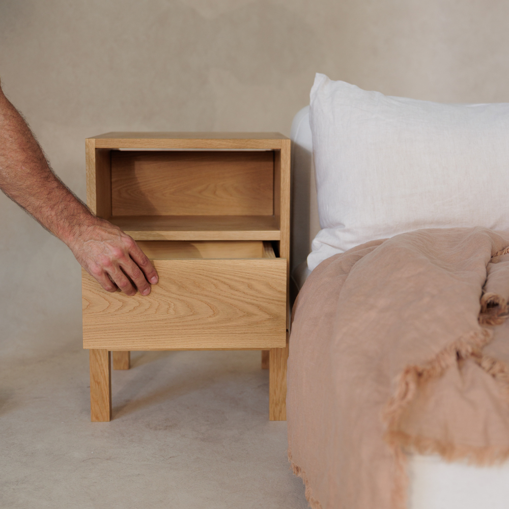 Wooden bedside table with a hand opening the drawer, next to a bed with a beige wall in the background