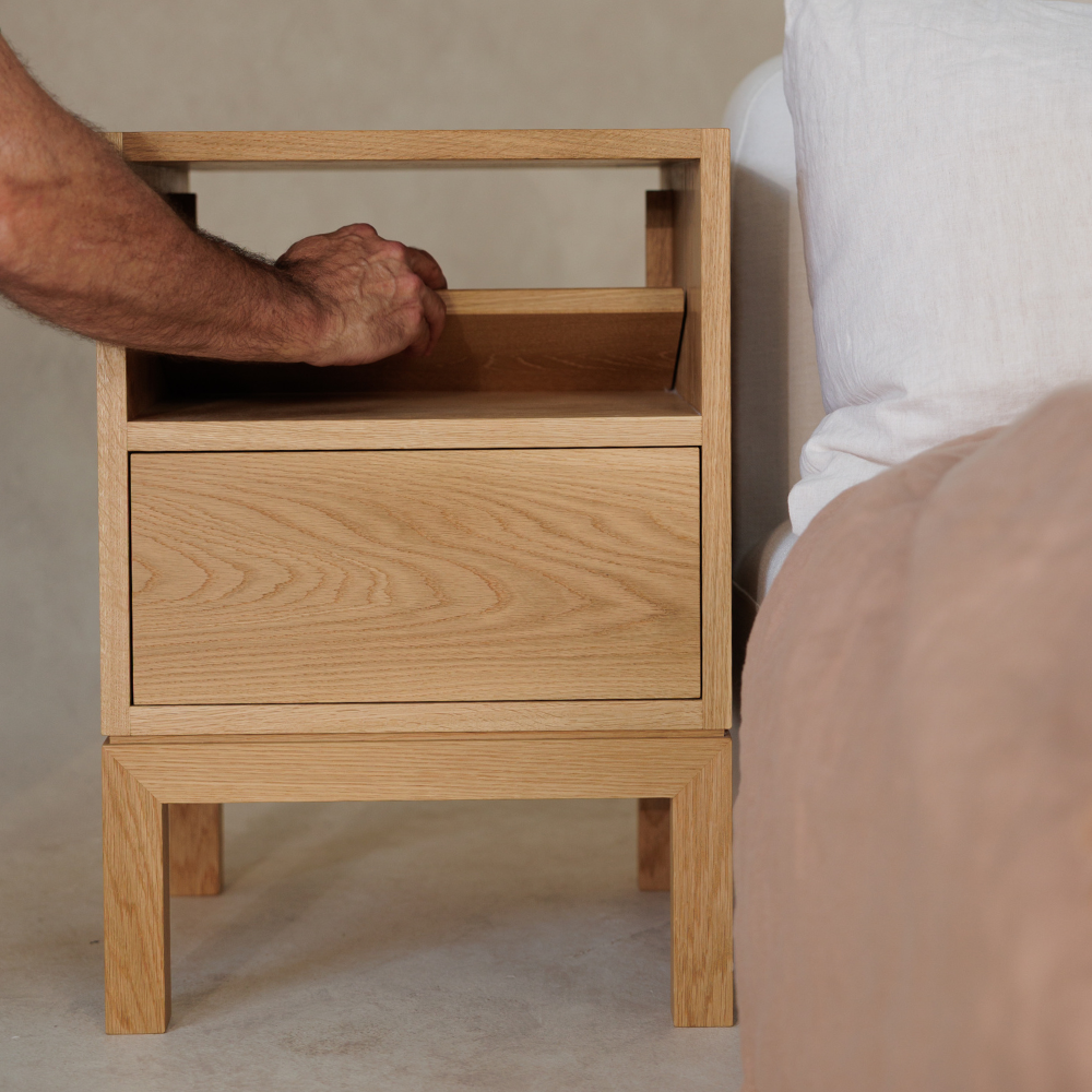 Wooden bedside table next to a bed with a hand opening the back panel