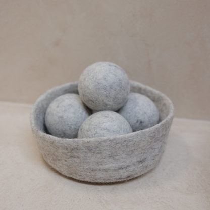 Grey felted dryer balls in a matching bowl on a beige surface.
