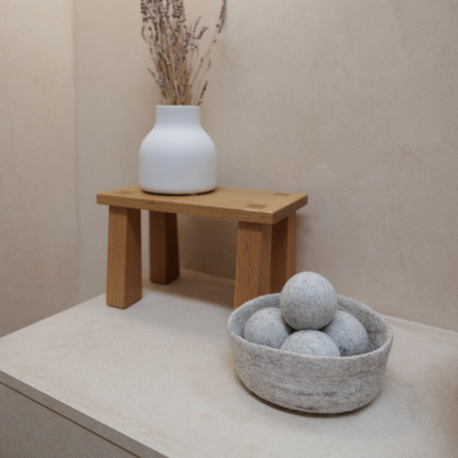 Wooden stool with a white vase and a grey bowl with wool balls on a beige surface.