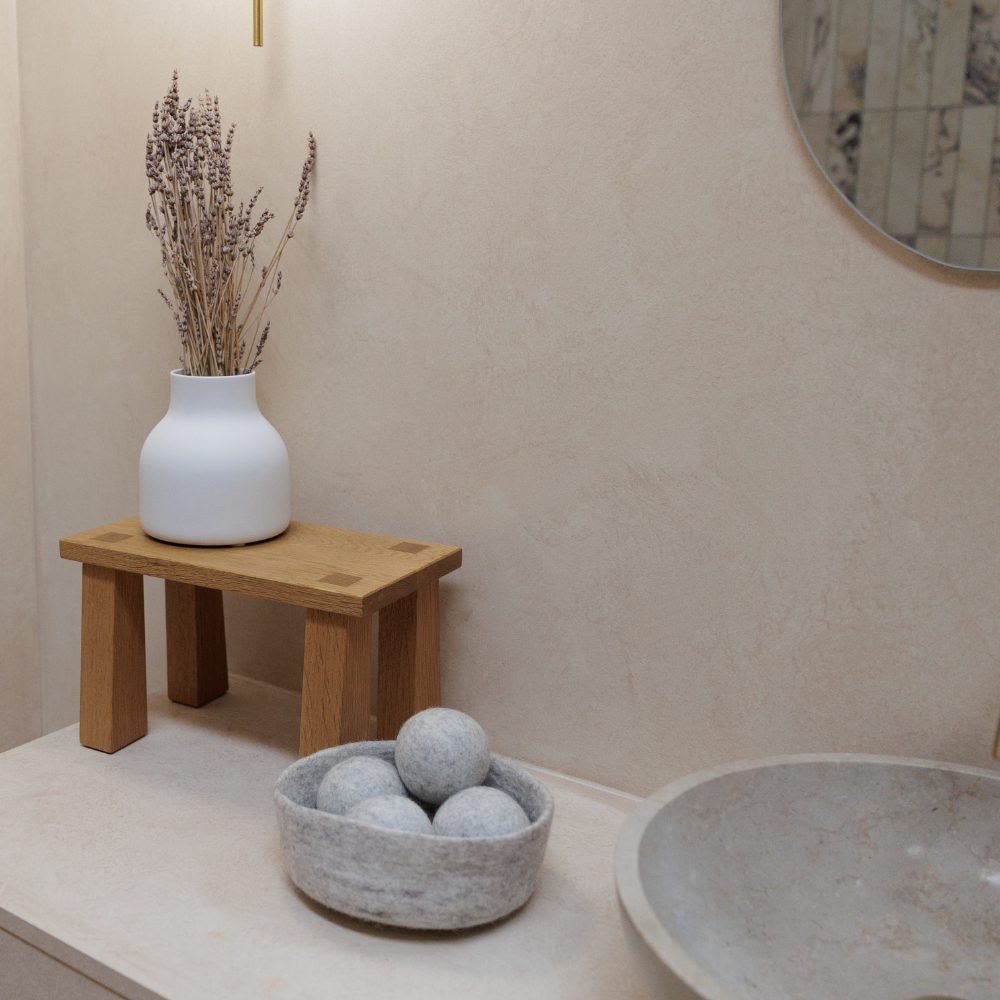Decorative elements including a white vase with dried lavender on a wooden stool, a felt bowl holding dryer balls, and a round mirror in a bathroom setting.