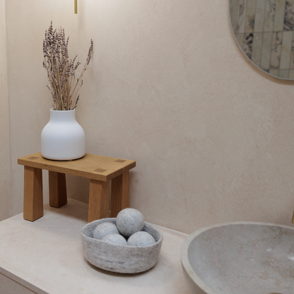 Decorative elements including a white vase with dried lavender on a wooden stool, a felt bowl holding dryer balls, and a round mirror in a bathroom setting.