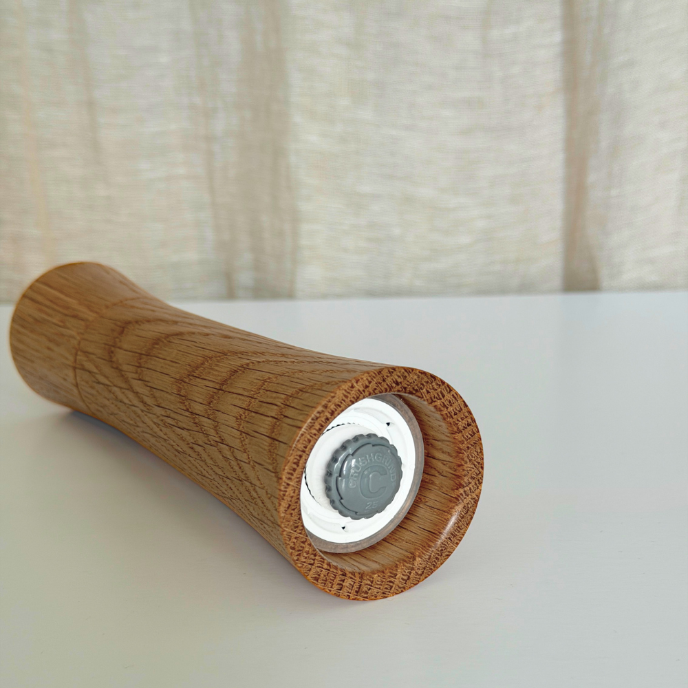 A wood pepper mill resting on a white surface with a beige fabric curtain in the background. Premium Crushgrind mechanism is featured