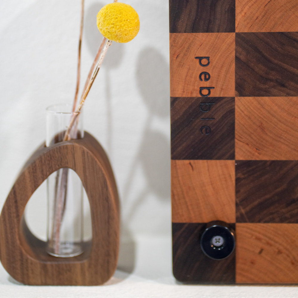 Wooden vase with a yellow flower and checkered wooden board with 'pebble' branding.