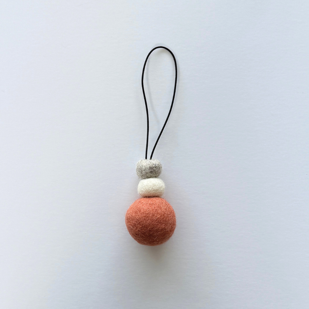Hand-felted wool hanging diffuser in soft sunset pink with neutral accent beads and black loop, photographed on a light background. Ideal for essential oils in car or home.