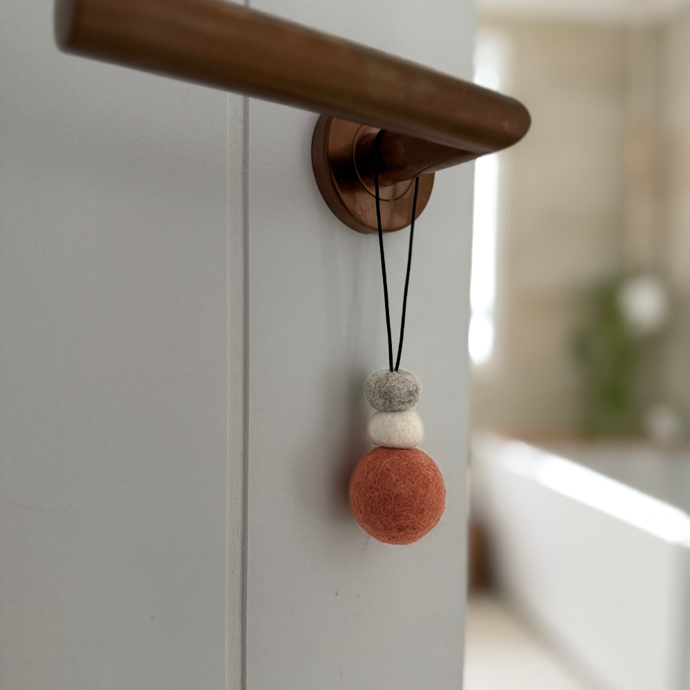 Hand-felted wool hanging diffuser in soft sunset pink with neutral accent beads and black loop, photographed on a door handle. Ideal for essential oils in car or home.