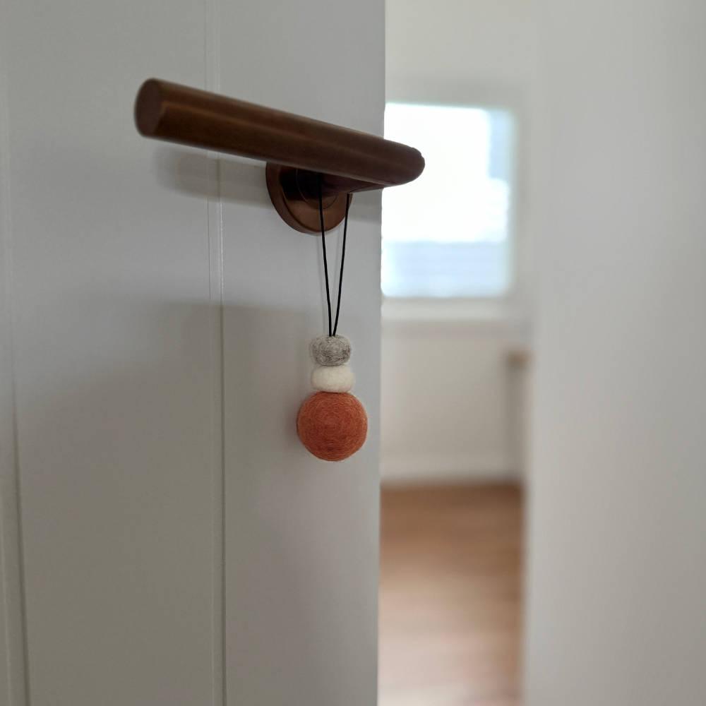 Hand-felted wool hanging diffuser in soft sunset pink with neutral accent beads and black loop, hanging from a door handle. Ideal for essential oils in car or home.