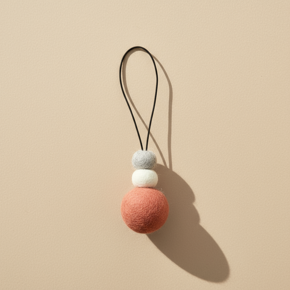 Hand-felted wool hanging diffuser in soft sunset pink with neutral accent beads and black loop, photographed on a light background. Ideal for essential oils in car or home.