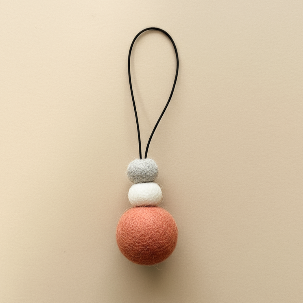 Hand-felted wool hanging diffuser in soft sunset pink with neutral accent beads and black loop, photographed on a light background. Ideal for essential oils in car or home.