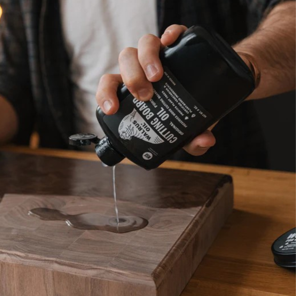 Person pouring Walrus Oil Cutting Board Oil from a black bottle onto a wooden chopping board.