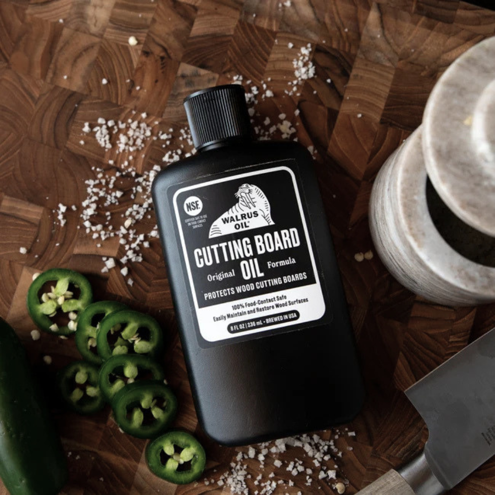 A bottle of Walrus Oil Cutting Board Oil is displayed on a rustic wooden cutting board, surrounded by sliced jalapeños, coarse salt, and a knife.
