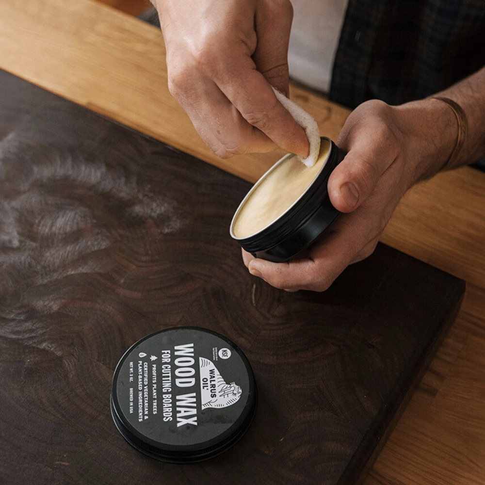 Person applying wood wax to a wooden surface with a container labelled 'Wood Wax for Cutting Boards'.