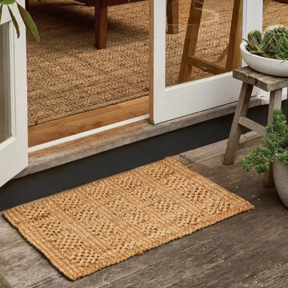 Armstrong rectangular natural jute doormat with a woven basketweave pattern, placed on a dark wood porch floor in front of white-framed glass doors.