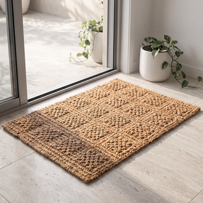 Armstrong rectangular woven jute doormat with a textured geometric pattern, placed on a light stone floor in front of a glass sliding door with potted plants nearby.