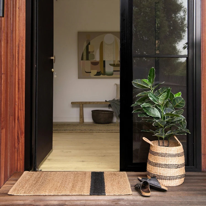 Baxter natural jute entryway doormat with a single black stripe down the centre, placed on a dark wood porch next to a pot plant and a pair of shoes, with an open black door revealing a minimalist interior.