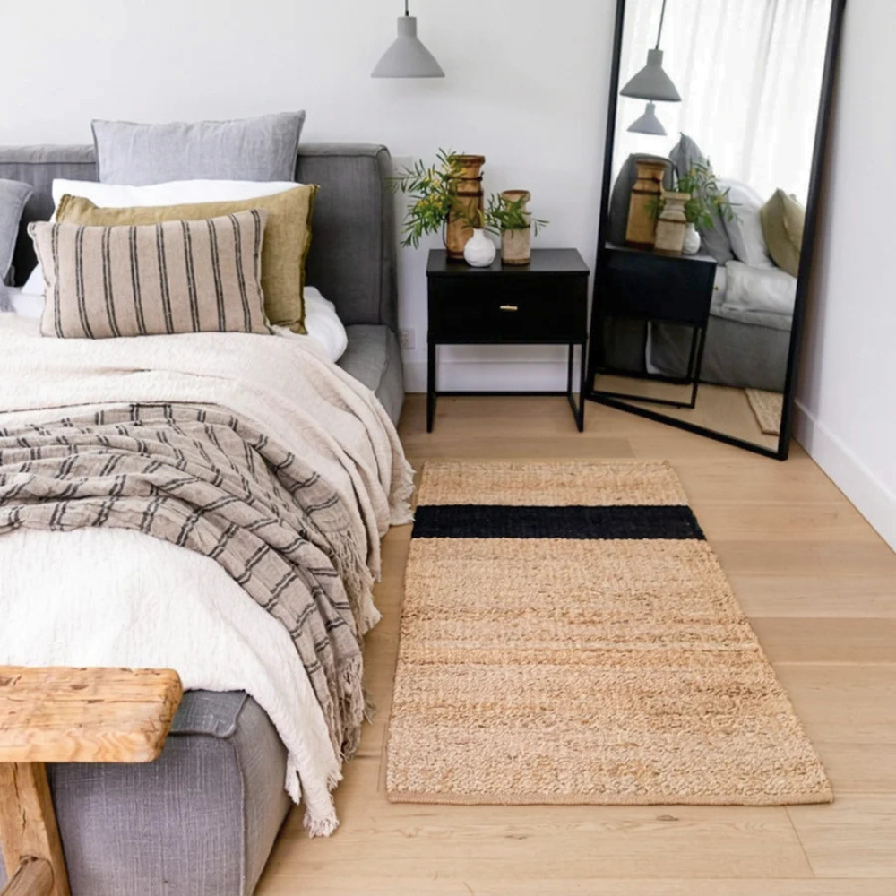 Baxter natural jute mat with a black stripe, styled on a light wood floor in a modern bedroom next to a grey bed and bedside table.