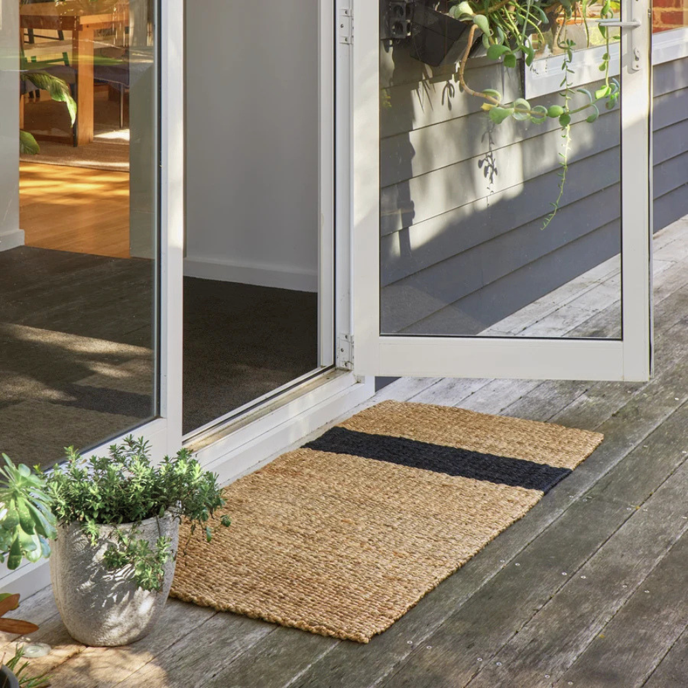 Baxter rectangular woven jute doormat with a black horizontal stripe, placed on a weathered wooden deck in front of white-framed glass patio doors.