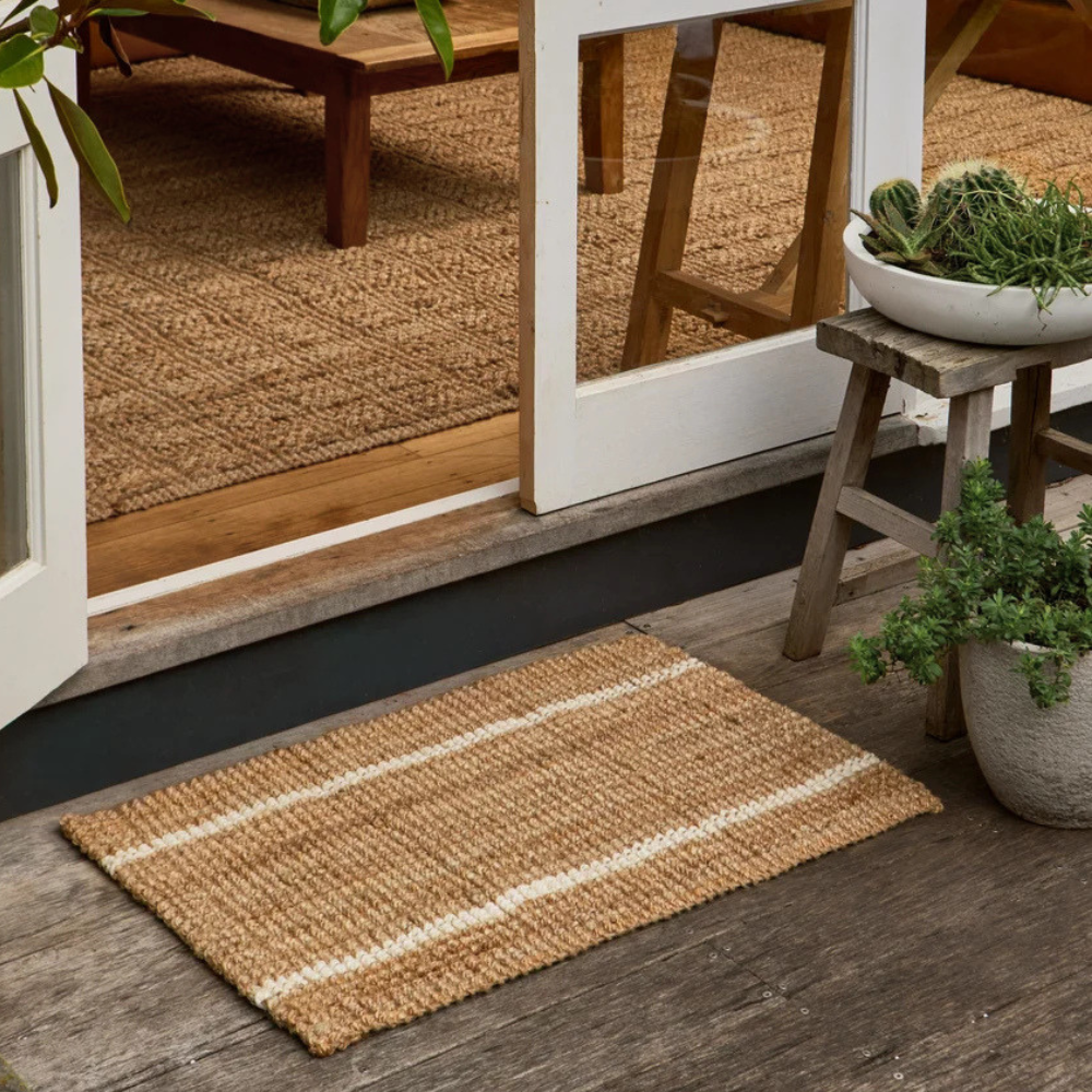 Robinson natural woven jute doormat with two cream-coloured stripes, placed on a weathered wooden deck in front of a glass door. A rustic wooden stool with a potted plant sits to the right.