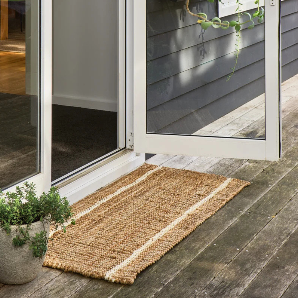 Robinson natural woven jute doormat with two cream-coloured stripes, placed on a weathered wooden deck in front of an open white-framed glass door.